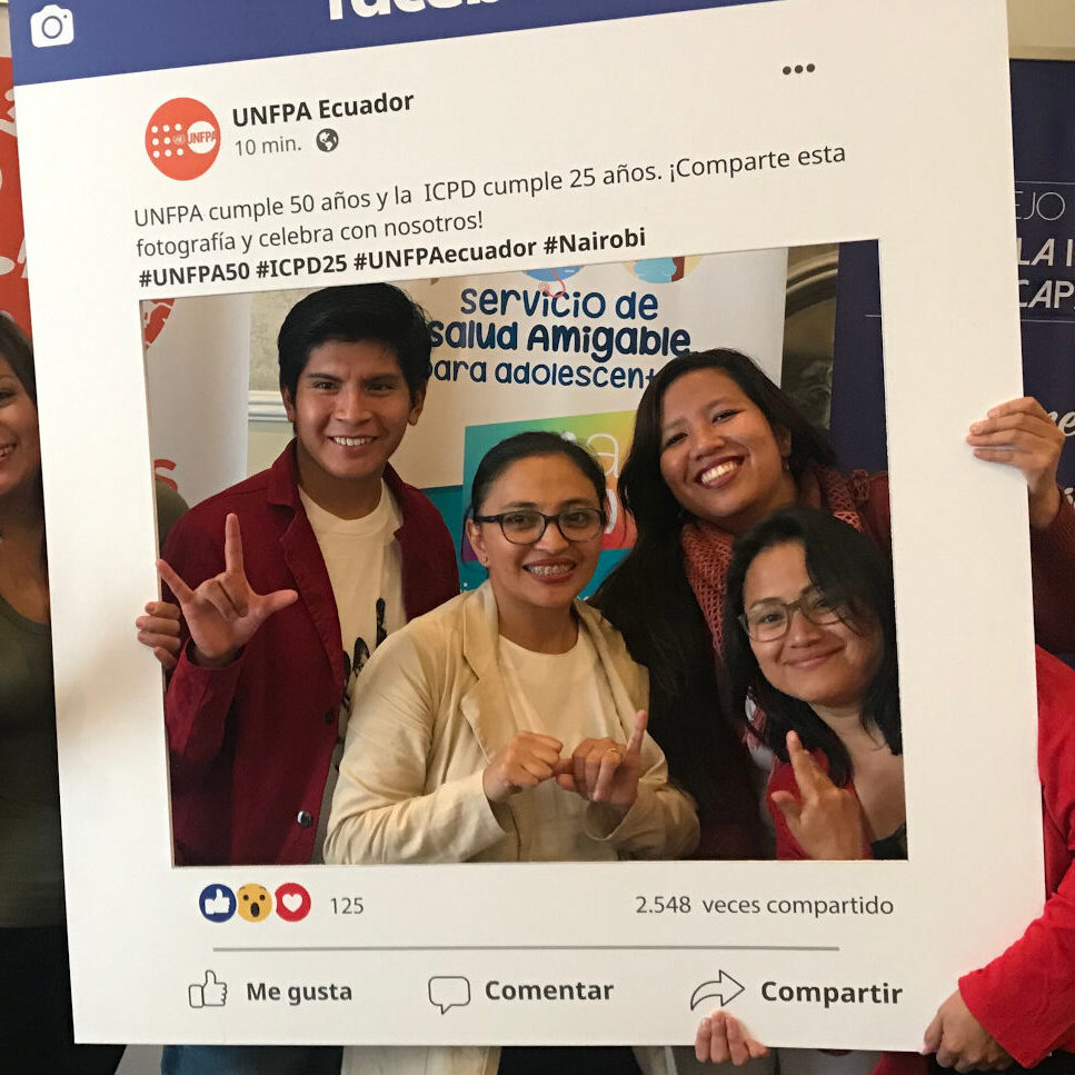 Fotografía de 4 jóvenes (un hombre y tres mujeres) sonriendo y haciendo distintas señas con las manos, mientras sostienen un marco de fotos grande que simula una publicación de las redes sociales de UNFPA Ecuador por el 50 aniversario de UNFPA y los 25 años de la IPCD.